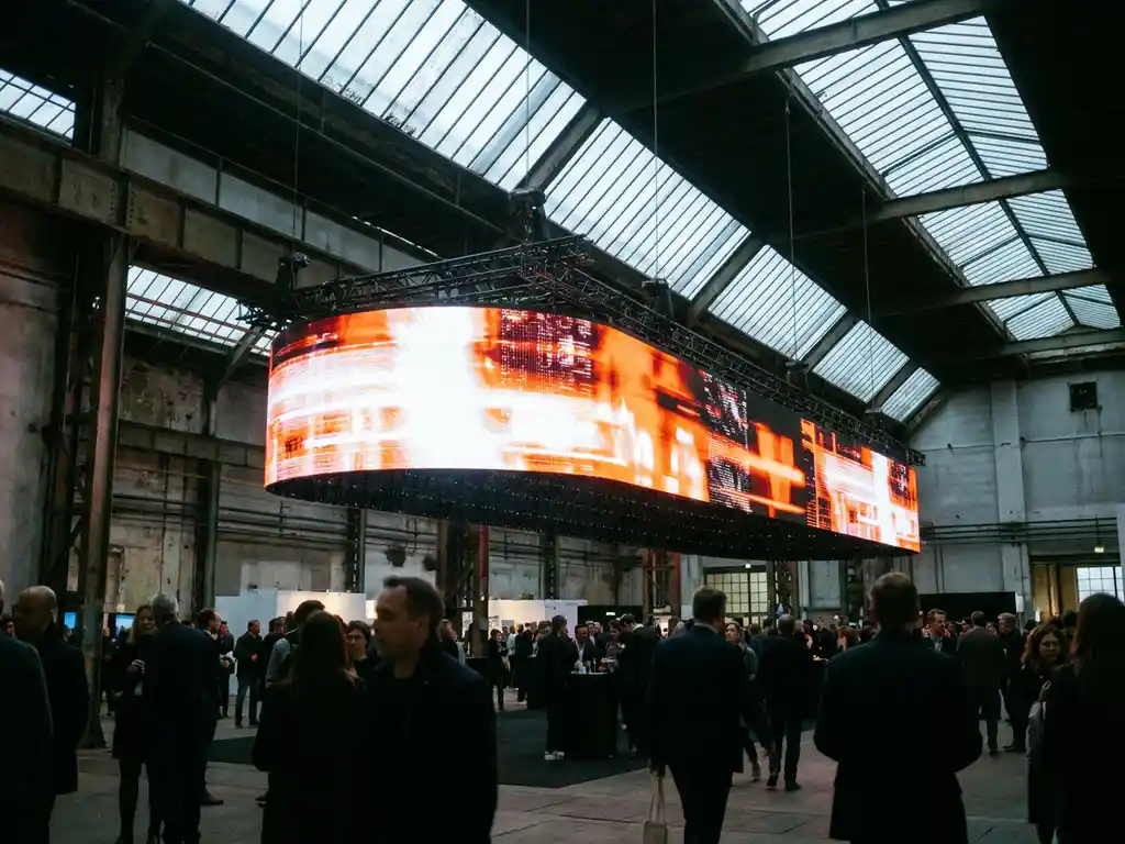 Großes leuchtendes LED-Banner in einer weitläufigen Messehalle mit Oberlichtfenstern und dramatischem Kontrast zwischen warmem und kühlem Licht.
