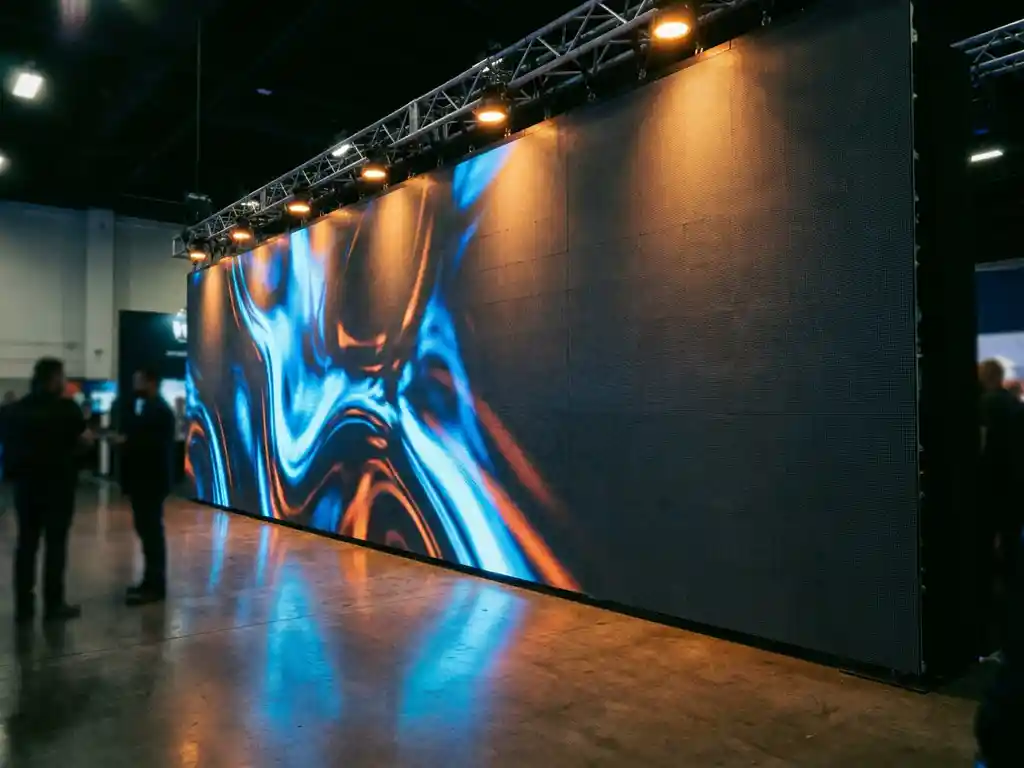 led-wand-messe-stand-display.jpg Große LED-Wand mit leuchtendem Content in einem modernen Messestand, beleuchtet von warmem Bernstein-Bühnenlicht auf poliertem Betonboden.